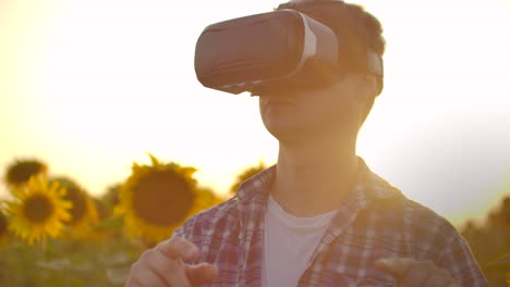 A-young-farmer-manager-in-plaid-shirt-and-jeans-uses-VR-glasses-on-the-field-with-sunflowers-for-scientific-article.-These-are-modern-technologies-in-summer-evening.